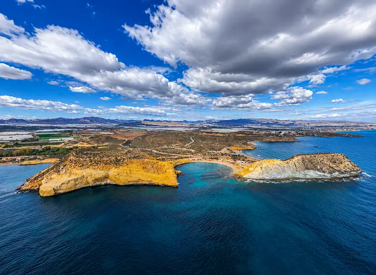 Las Cuatro Calas desde el aire, con entrantes de roca dorada entre agua turquesa y nubes de tormenta al fondo