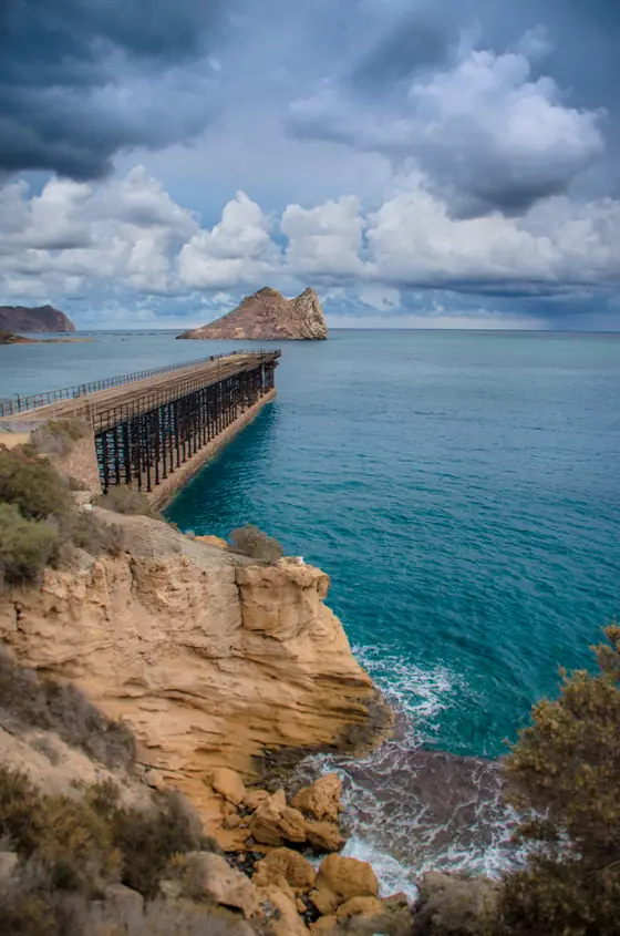 El embarcadero del Hornillo visto desde el acantilado, con el agua turquesa y nubes de tormenta sobre la isla del Fraile
