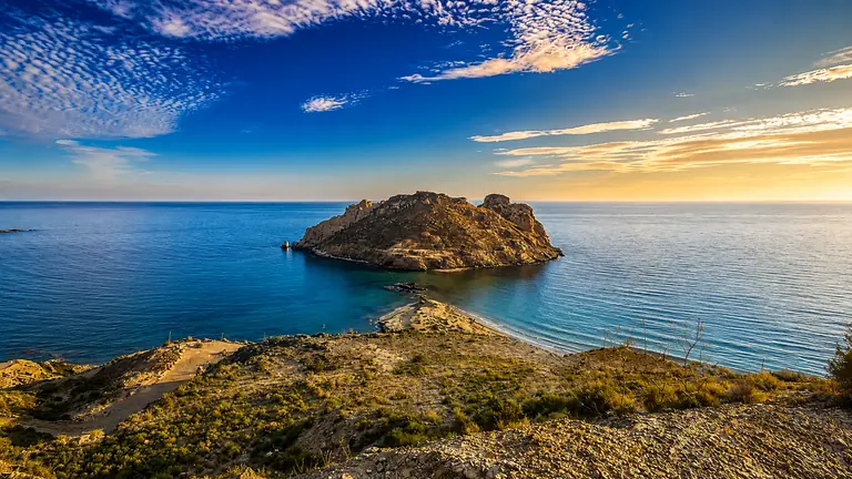 La isla del Fraile al atardecer, unida a la costa por una lengua de arena, bajo un cielo de nubes altas