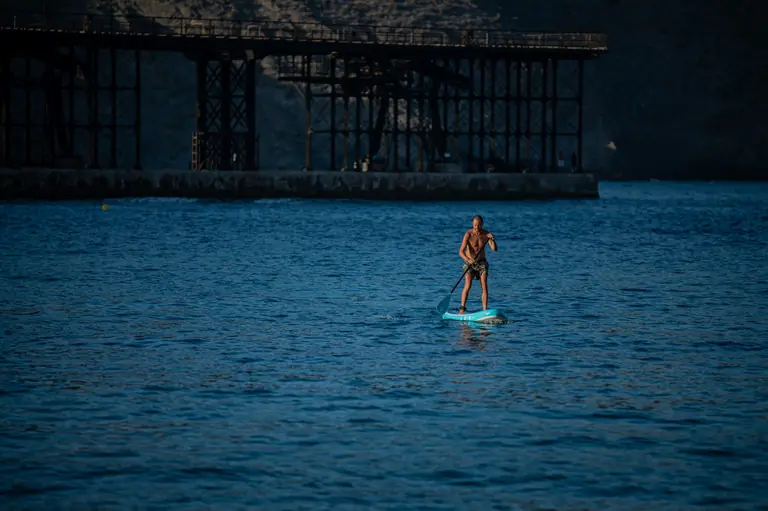 Una persona rema de pie sobre una tabla de paddle surf a contraluz, con la estructura del embarcadero del Hornillo detrás