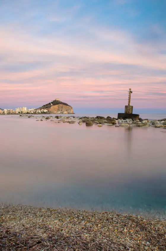 Una playa de cantos rodados al anochecer, con el mar en calma y el castillo de San Juan iluminado al fondo