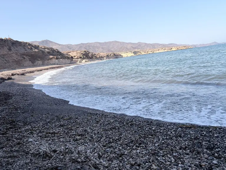 La curva de Playa Larga, con la orilla de grava oscura, la línea de espuma y la sierra al fondo